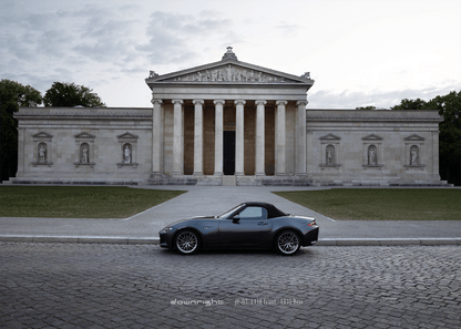 Grey Mazda MX-5 ND with shinny wheels from downright parked on a cobblestone street in front of a classical building