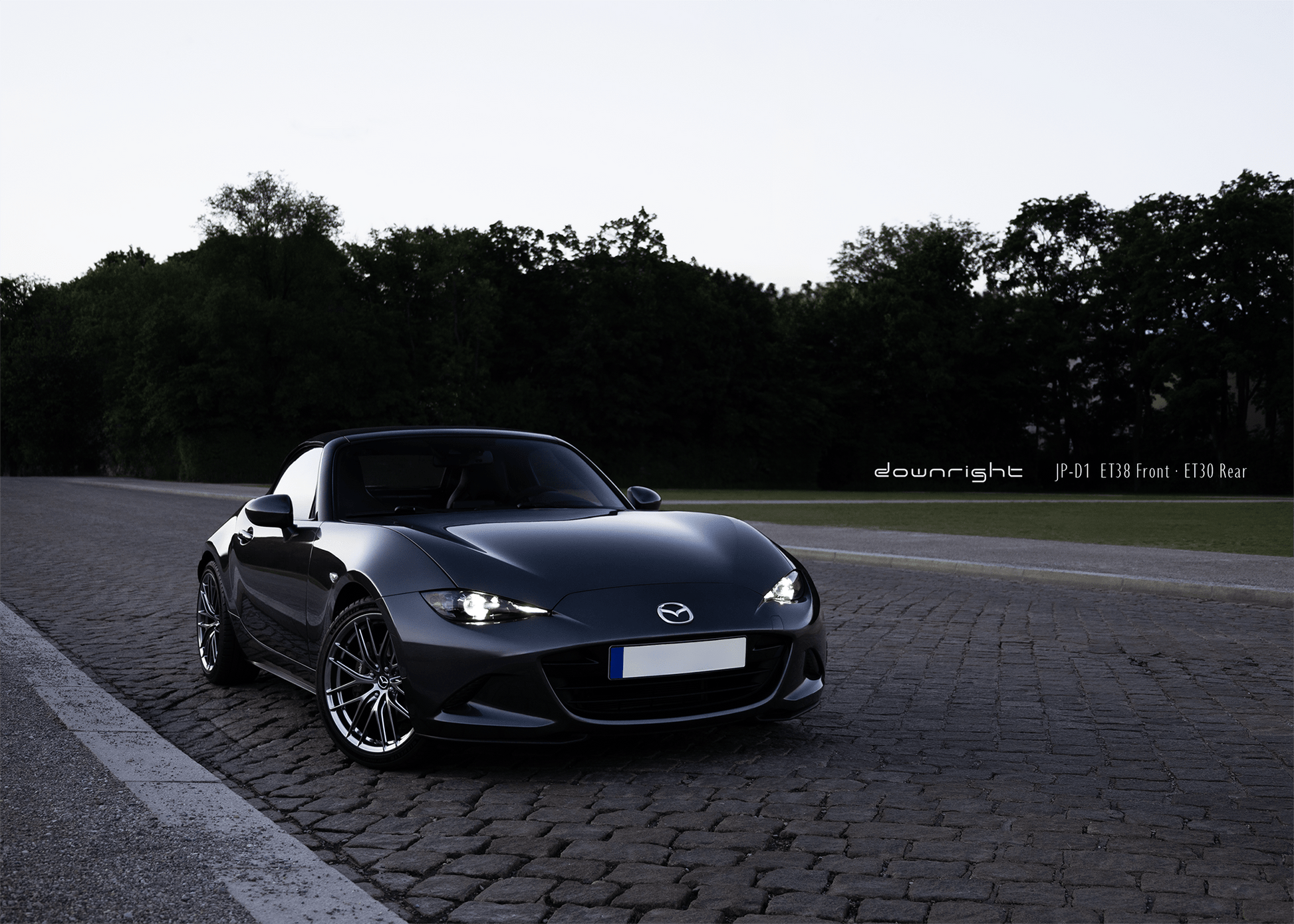 Grey Mazda MX-5 ND with shinny wheels from downright on a road with trees in the background