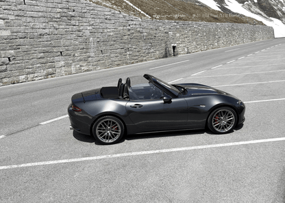 Grey Mazda MX-5 ND with shinny wheels from downright on a mountain road of Grossglockner with stone wall and snow-capped mountains of Alps in the background